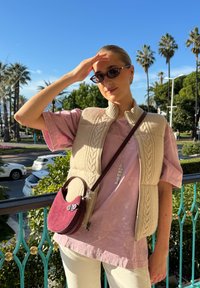 Jeune femme portant des lunettes de soleil, un gilet en tricot beige, une chemise rose et un pantalon blanc, se protégeant les yeux tout en se tenant sur un balcon avec des palmiers en arrière-plan.