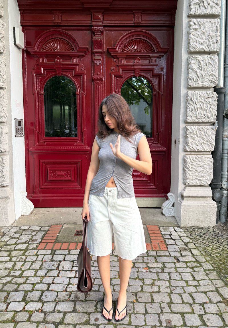 Jeune femme en haut gris sans manches et short blanc se tenant dans une rue pavée devant de grandes portes doubles rouges foncées détaillées, tenant un sac à main marron.
