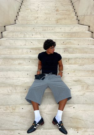 Camisa negra ajustada, pantalones cortos grises claros hasta la rodilla, calcetines blancos, mocasines negros con una tira decorativa. Sentado en escalones de piedra beige.