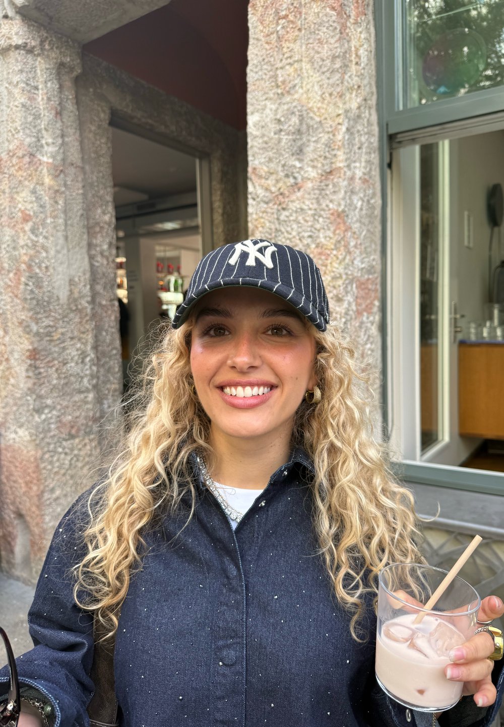 Ung kvinna med lockigt blont hår och NY-keps som ler, håller i ett glas med is och en ljus dryck, stående utomhus vid en stenmur.