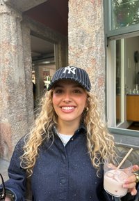 Ung kvinna med lockigt blont hår och NY-keps som ler, håller i ett glas med is och en ljus dryck, stående utomhus vid en stenmur.