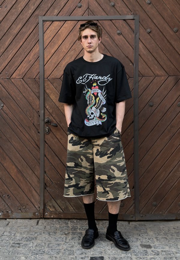 Mladý muž stojí s rukami v kapsách, nosí čierne tričko Ed Hardy, maskáčové šortky, čierne ponožky a topánky na pozadí drevených dverí.