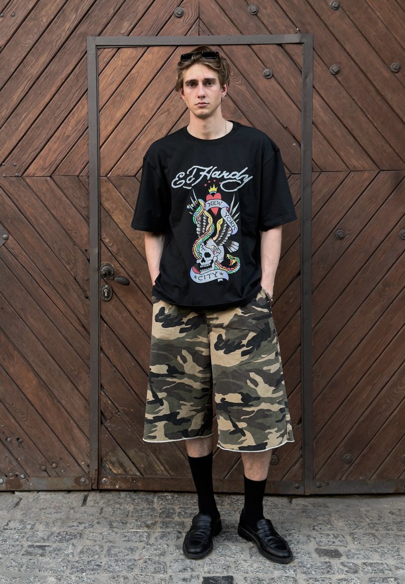 Jeune homme debout, mains dans les poches, portant un t-shirt noir Ed Hardy, un short camouflage, des chaussettes noires et des chaussures devant une porte en bois.