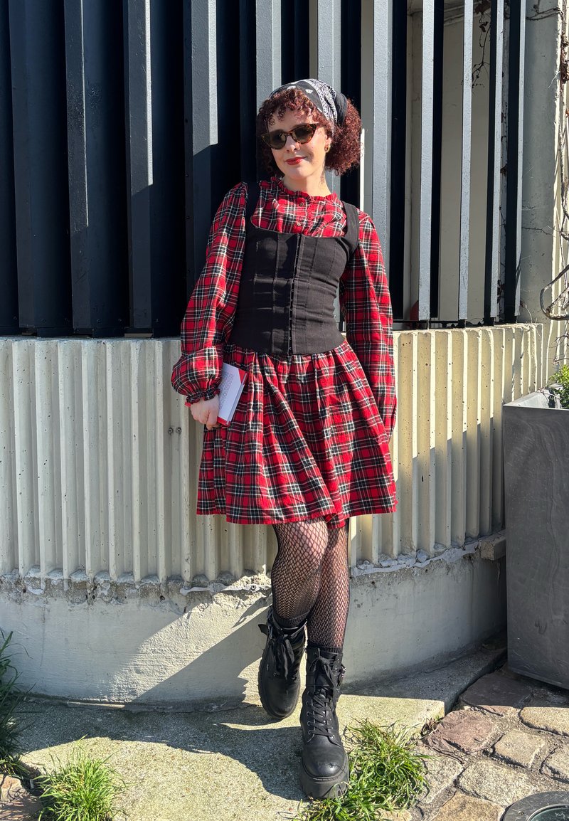 Femme portant une robe à carreaux rouges, un corset noir, des collants résille et des bottes noires, debout contre un mur en béton côtelé en plein soleil.