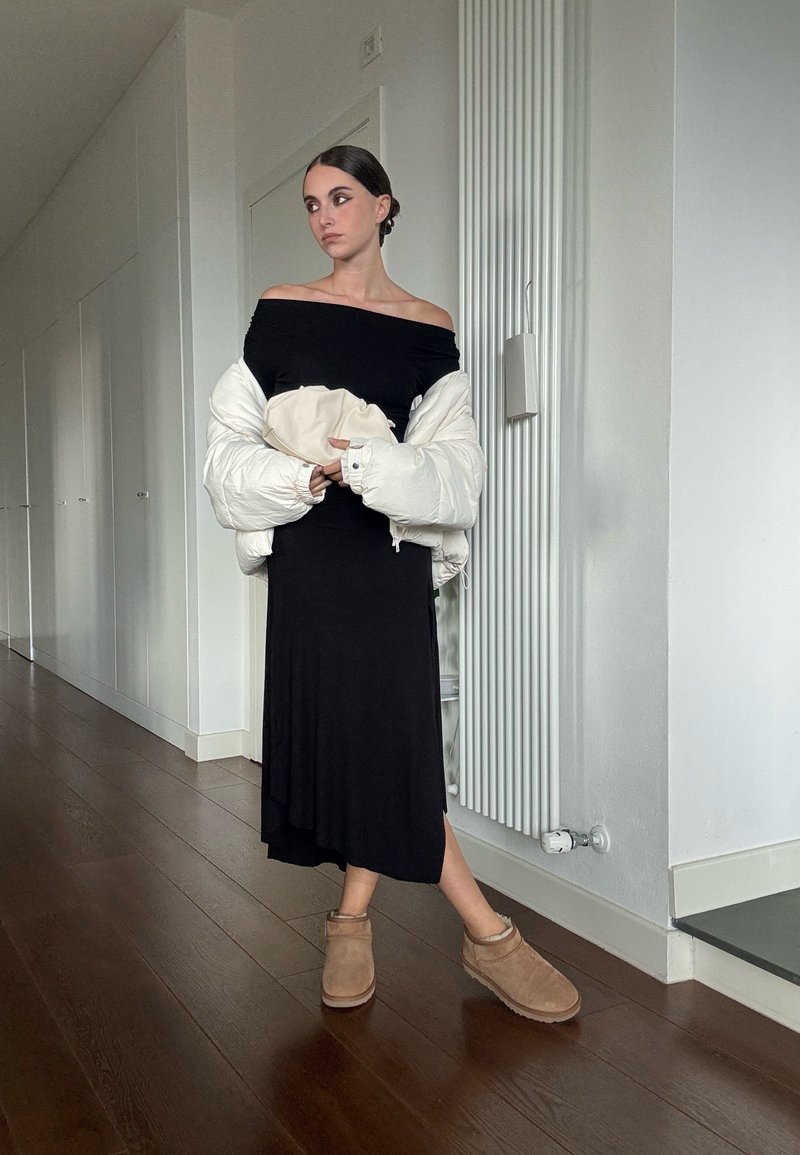 Γυναίκα με μαύρο off-shoulder φόρεμα, λευκή πούφ φανέλα ριγμένη στους ώμους, κρατώντας τσάντα σε λευκό χρώμα, φορώντας καφέ μπότες μέχρι τον αστράγαλο στο εσωτερικό.