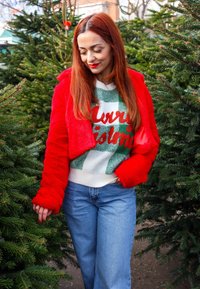 Γυναίκα με μακριά κόκκινα μαλλιά, ντυμένη με κόκκινο μπουφάν και Χριστουγεννιάτικο πουλόβερ, περπατάει ανάμεσα σε έλατα σε μια υπαίθρια αγορά δέντρων.