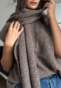 Brązowy, teksturowany szal owinięty wokół szyi, uzupełniony srebrnymi pierścieniami na palcach. Tkanina wykazuje subtelne odcienie koloru i wzoru.