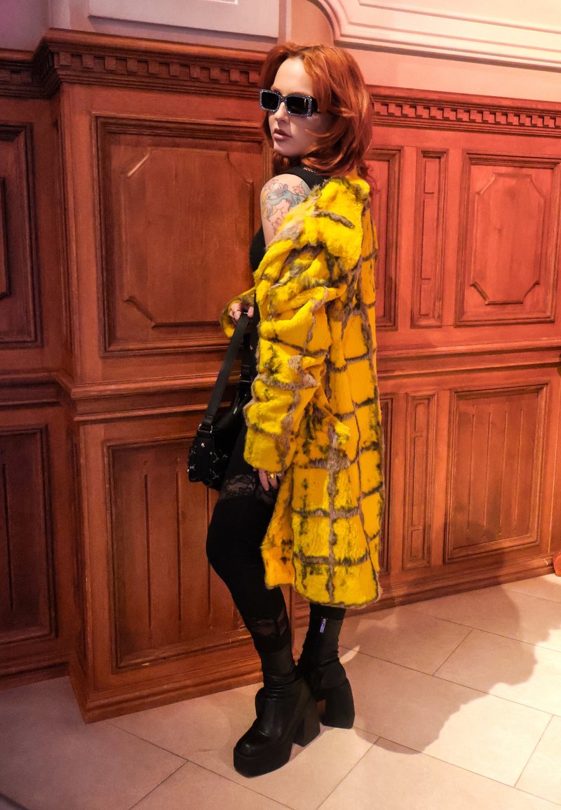 Femme aux cheveux roux portant des lunettes de soleil noires carrées, un manteau en fausse fourrure jaune à carreaux, une robe noire, des bottes à plateforme et tenant un sac à main noir.