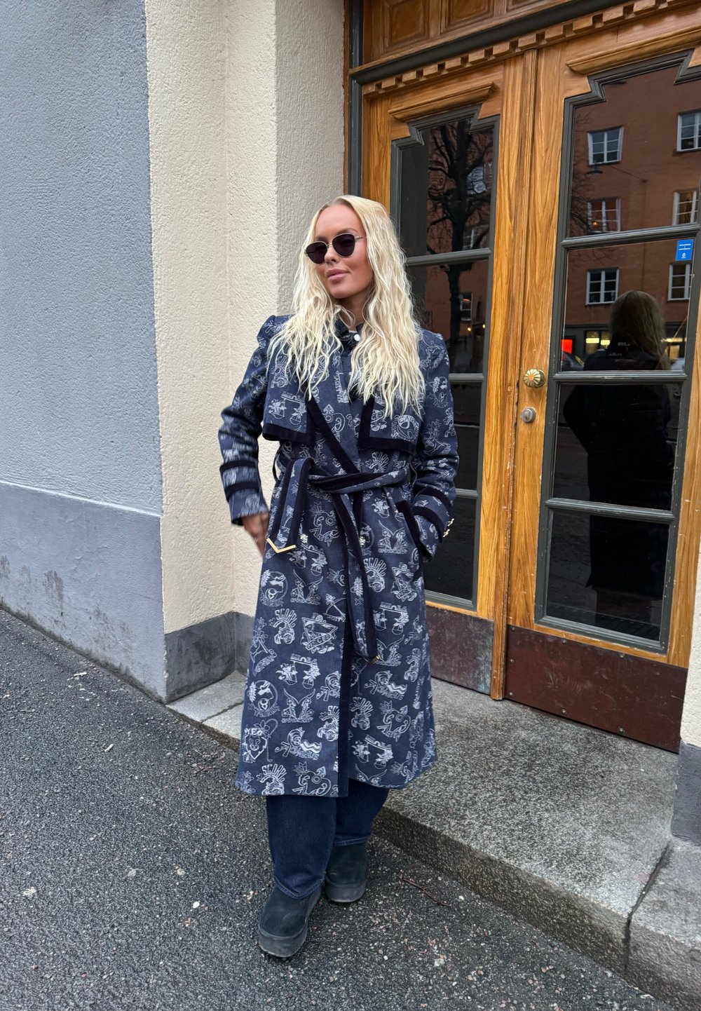 Μπλε παλτό με σχέδιο, με ζώνη και μακριά μανίκια, φορεμένο πάνω από σκούρα τζιν και μαύρες μπότες, έξω κοντά σε ξύλινη πόρτα.