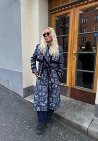 Μπλε παλτό με σχέδιο, με ζώνη και μακριά μανίκια, φορεμένο πάνω από σκούρα τζιν και μαύρες μπότες, έξω κοντά σε ξύλινη πόρτα.