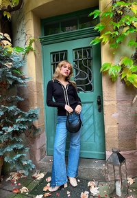 Cardigan nero con bordatura bianca, jeans svasati blu chiaro, borsa circolare nera, in piedi accanto a una porta verde con una grata in metallo decorata.