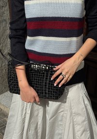 Black woven handbag with a rectangular shape and slim handles, held by a hand wearing rings, paired with a striped knit sweater and a pleated skirt.