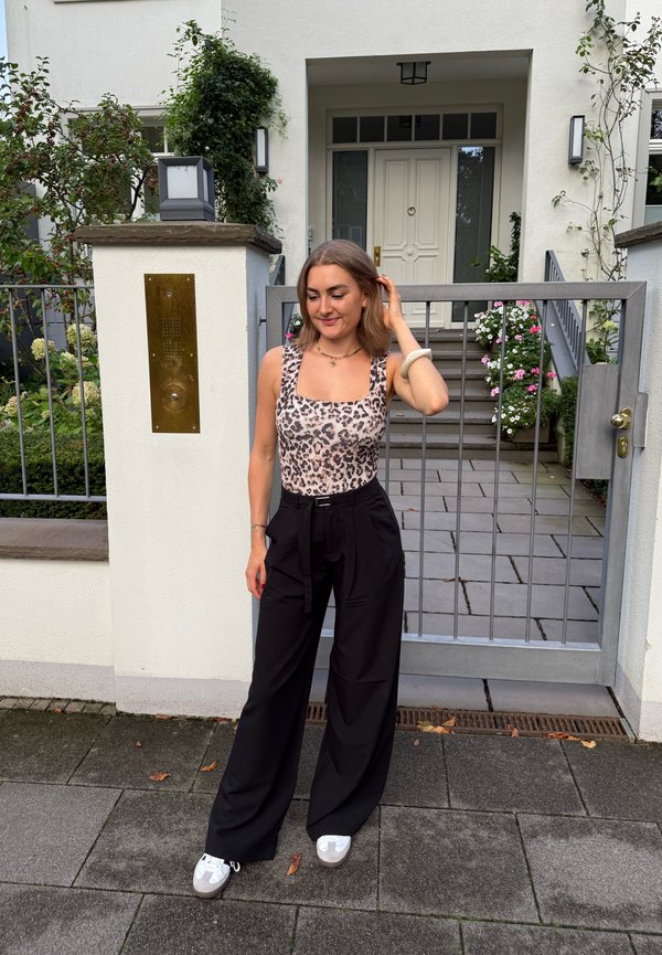 Bodysuit z leopardjim vzorom v kombinaciji z črnimi širokimi hlačami z visoko pasom in belimi supergami. Stoji ob sivi ogradi s rastlinami v ozadju.