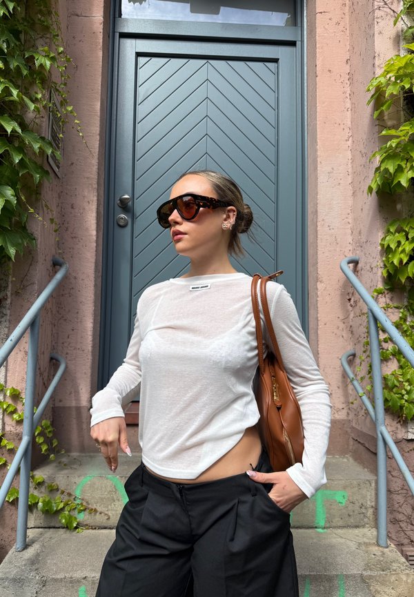 Donna con occhiali da sole, top bianco a maniche lunghe e pantaloni neri, in piedi sulle scale accanto a una porta blu con edera verde sui muri.