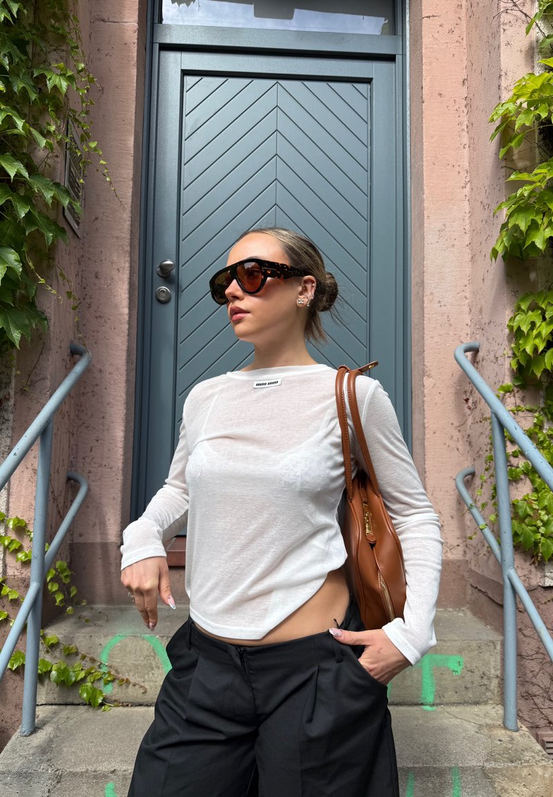 Femme portant des lunettes de soleil, un haut blanc à manches longues et un pantalon noir, debout dans des escaliers près d'une porte bleue avec du lierre vert sur les murs.