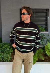 Sweater listrado em preto e verde com colarinho e punhos canelados, apresentando bordados em ouro. Calças cargo bege com bolsos laterais e etiqueta de marca.
