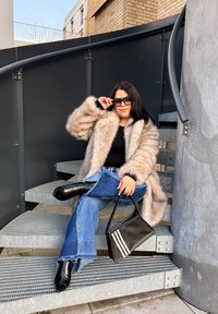 Femme en manteau en fausse fourrure beige et lunettes de soleil, assise jambes croisées sur des escaliers métalliques, tenant un sac à main noir avec des rayures blanches.