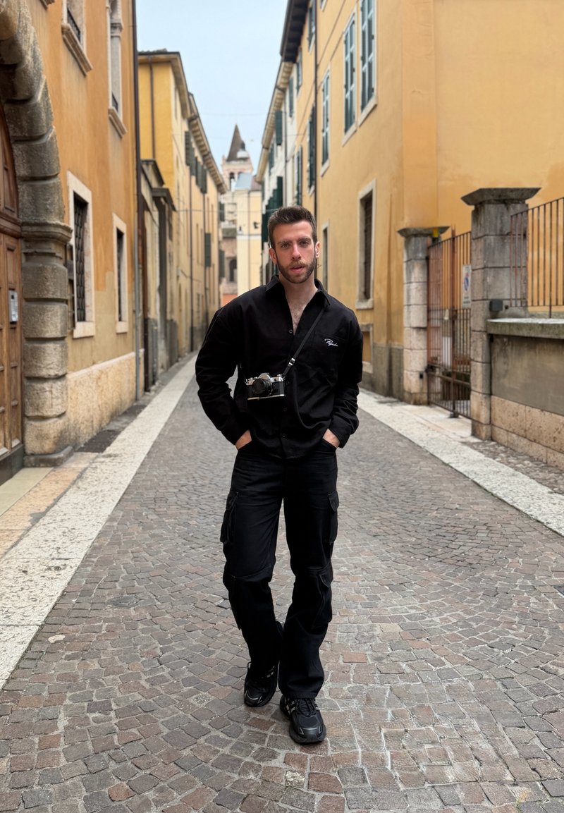 Νέος άνδρας με μαύρη στολή και φωτογραφική μηχανή γύρω από το λαιμό στέκεται σε λιθόστρωτο δρόμο ανάμεσα σε κίτρινα κτίρια.