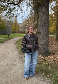Oversized brun läderjacka med vida ärmar, kombinerad med ljusblå baggy-jeans och en svart handväska, i en parkmiljö med träd.