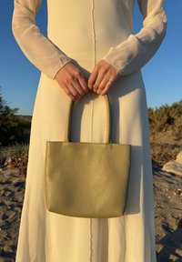 Persoană îmbrăcată într-o rochie lungă bej, ținând o geantă mică, texturată, de culoare verde măslin, în aer liber, la apus.