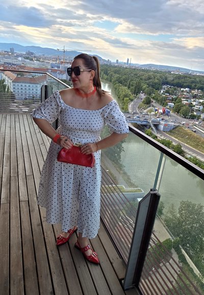 Vestido blanco sin mangas con lunares negros, bolso rojo y zapatos rojos a juego. Horizonte claro al fondo. Suelo de madera.