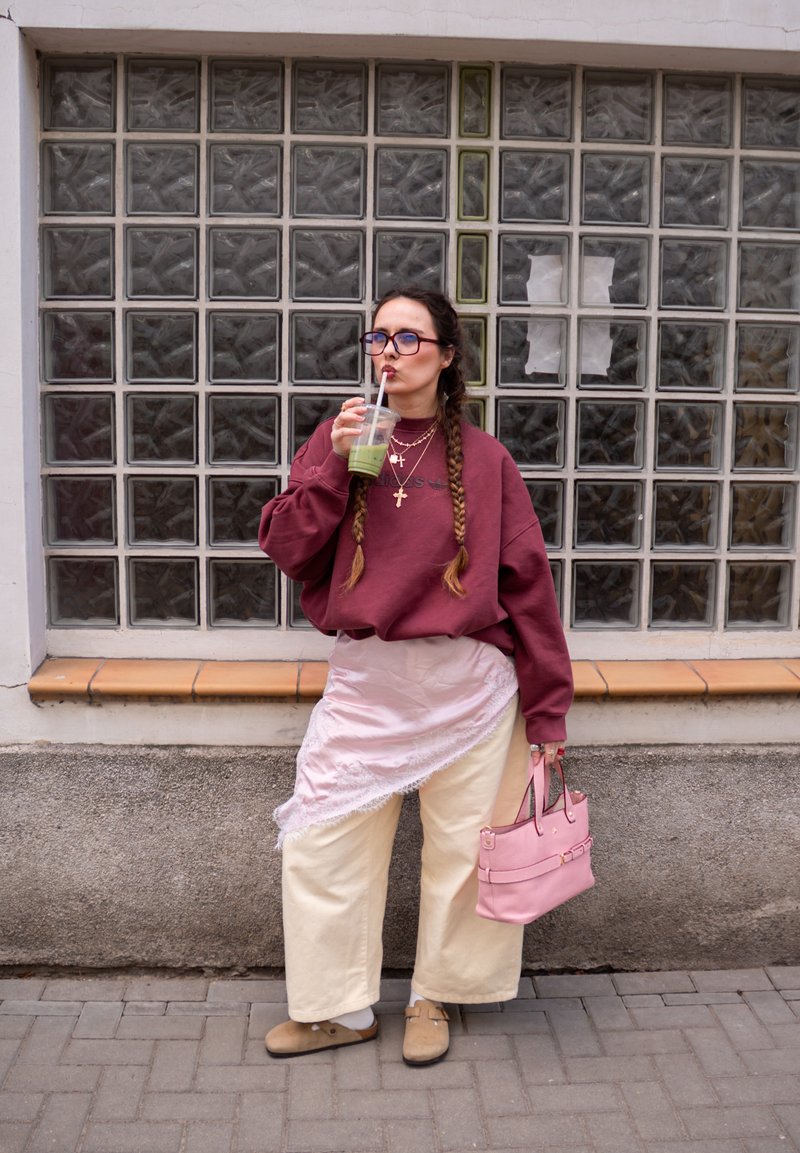 Žena nosící burgundský sweatshirt a krémové kalhoty, držící růžovou kabelku, pije zelený nápoj na pozadí zdi ze skleněných bloků.