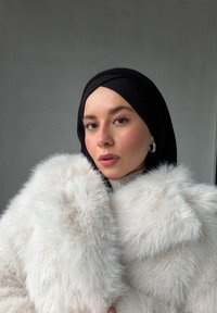 Abrigo de piel sintética blanca con cuello grande, llevado sobre un pañuelo negro de punto. Tono de piel claro, maquillaje sutil y pendientes de aro visibles.