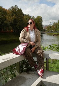 Μπεζ παλτό, άσπρο πουκάμισο με κουμπιά, παντελόνια με λεοπάρ σχέδιο, γυαλιστερή μπορντό τσάντα, κόκκινα και άσπρα αθλητικά παπούτσια, καθισμένος κοντά σε ποτάμι με πράσινο τοπίο.