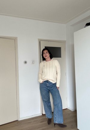 Mujer de pie en el interior, con un suéter crema de punto trenzado, vaqueros azules de pierna ancha y botines oscuros con tacón, frente a una pared blanca lisa.