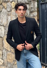 Černý blazer s jednou náprsní kapsou, zdobenou zlatým emblémem, nošený přes černou košili a světle modré džíny, umístěný na pozadí kamenné zdi.