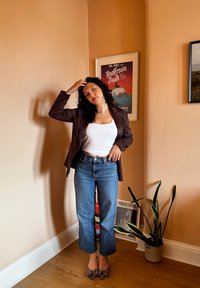 Smeđa blazerica preko bijelog majicama bez rukava, u kombinaciji sa svijetlim skraćenim trapericama i potpeticama sa leopard uzorkom. U pozadini saksijska biljka i plakati.