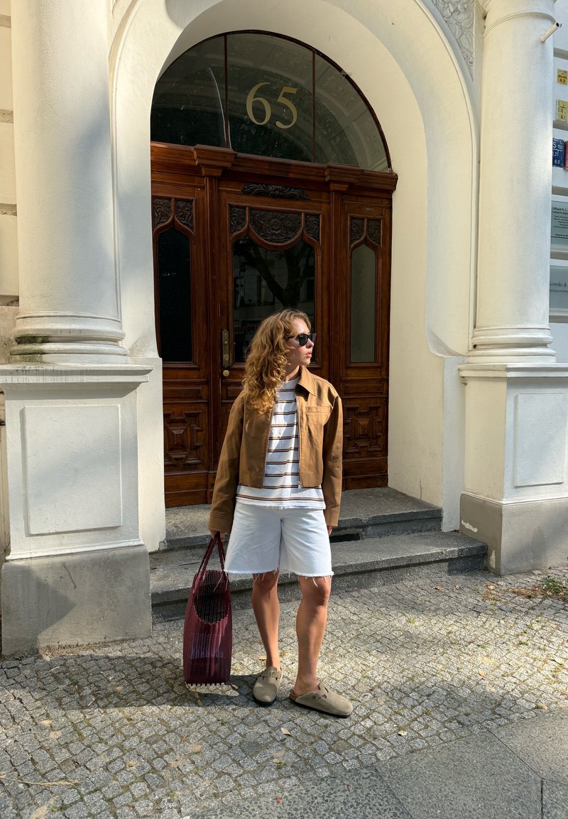 Woman in sunglasses, brown jacket, white shorts stands on cobblestone steps holding a red bag outside a building numbered 65 with wooden doors.