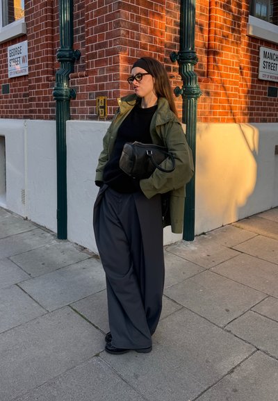 Chaqueta verde sobre un suéter negro, combinada con pantalones grises holgados. Bolso negro con acabado texturizado. De pie sobre una acera urbana.