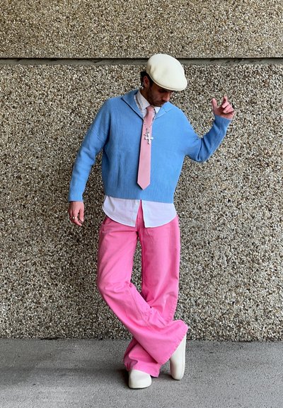Suéter azul claro acanalado, camisa blanca, corbata rosa, pantalones rosa de pierna amplia y zapatos blanco sucio. Collar cruzado notable y gorra beige.