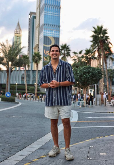 Camisa de rayas azul marino de mangas cortas, pantalones cortos beige y zapatillas ligeras; de pie en un camino pavimentado con edificios y palmeras de fondo.