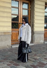 Chlupatý světle šedý kabát, pod ním bílá mikina, černé sportovní kalhoty s bílými pruhy a černá kabelka. Stojí poblíž dvojitých dřevěných dveří.