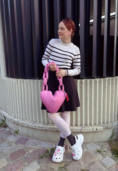 Bolso rosa en forma de corazón con un mango grueso, llevado por una persona con un suéter a rayas, falda negra, medias de lunares y zuecos blancos decorados.