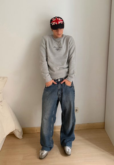 Sudadera gris con logo, vaqueros azules oversize y zapatillas deportivas grises. La gorra de béisbol presenta un diseño de la Union Jack en rojo y blanco.