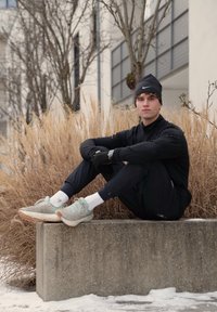 Jeune homme en tenue de sport noire et bonnet assis sur un bloc de béton en plein air, avec de l'herbe sèche et de la neige dans un environnement urbain hivernal.