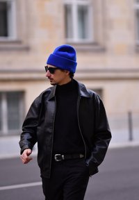 Άνδρας που περπατά σε αστική οδό φορώντας μαύρο δερμάτινο μπουφάν, μαύρο πουλόβερ με τσέπη, μπλε σκουφάκι, γυαλιά ηλίου και ζώνη με ασημένια πόρπη.