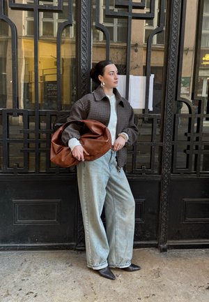 Mujer de pie frente a una reja decorativa de hierro negro, vestida con chaqueta marrón a cuadros, camisa blanca, vaqueros de pierna ancha azul claro, sosteniendo un bolso grande marrón.