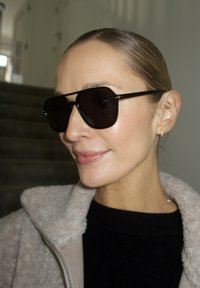 Lunettes de soleil noires surdimensionnées avec une monture épaisse et des accents dorés. La texture est lisse et les verres sont foncés, offrant une couverture complète.