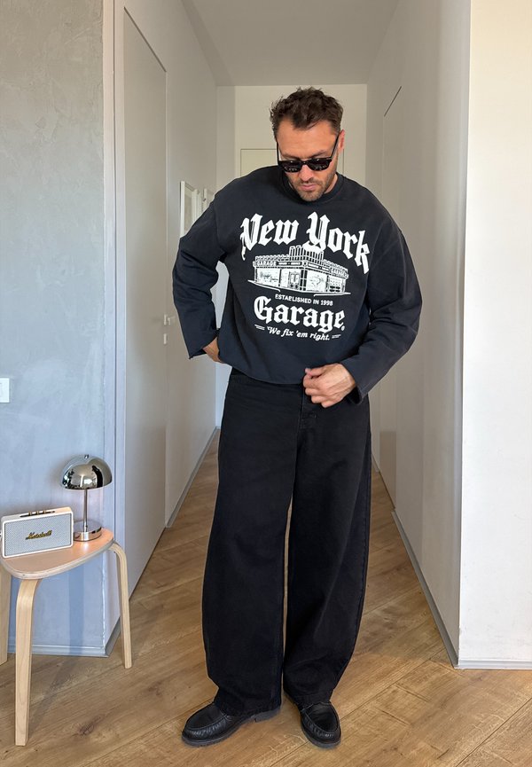 Hombre con gafas de sol negras, sudadera negra con estampado "New York Garage" y pantalones anchos negros, de pie en un pasillo ajustándose los pantalones.