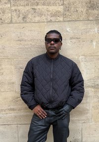 Blouson aviateur matelassé noir avec fermeture éclair avant, associé à un pantalon en cuir noir et un gant noir texturé. Des lunettes de soleil foncées complètent le look.