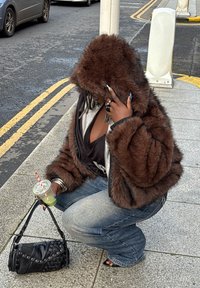 Personne portant une veste à capuche en fourrure brune accroupie sur le trottoir, tenant un verre avec une paille rayée et un sac à main noir, près de voitures garées.