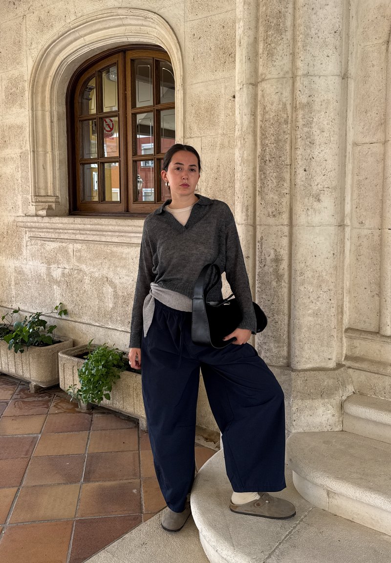 Pulôver cinza de malha com gola, sobre uma blusa clara, combinado com calças largas azuis-marinho e sapatos cinzentos de deslizar. Segura uma bolsa preta, em pé junto a uma arcada.