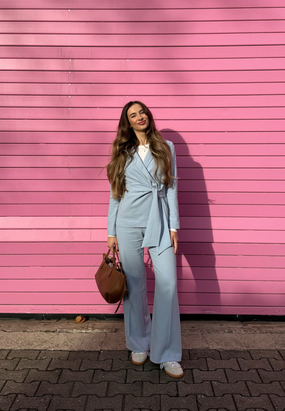 Completo azzurro chiaro con cintura in vita, pantaloni svasati, abbinato a una borsa marrone e scarpe da ginnastica bianche. Sfondo con listelli di legno rosa.