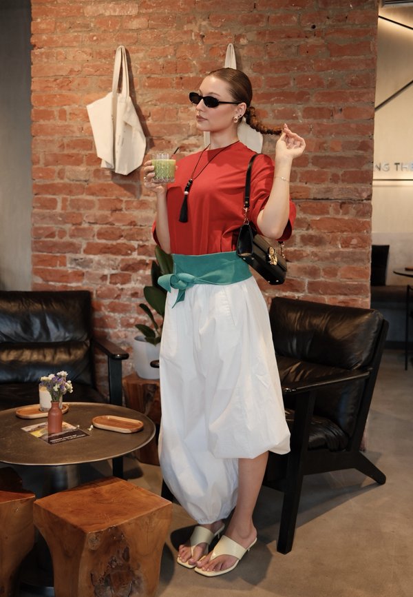 Femme en lunettes de soleil tenant une boisson verte, portant un haut rouge, un pantalon blanc avec une ceinture verte, debout dans un salon avec un mur en briques et des meubles en bois.