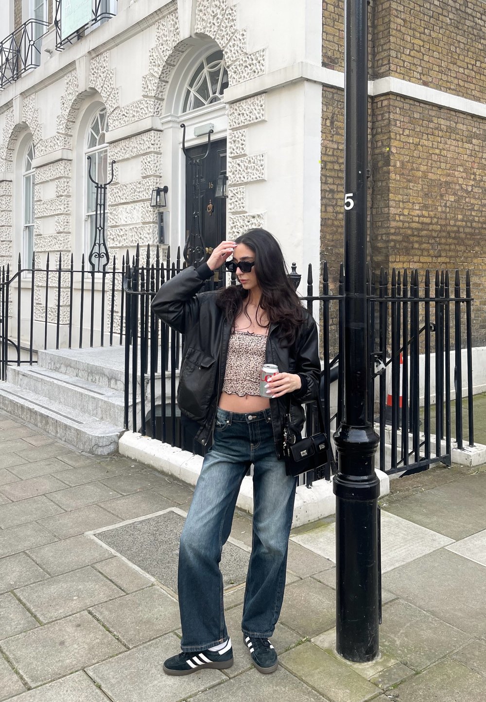 Μαύρο bomber jacket, τοπ σε λεοπάρ σχέδιο, φαρδιά μπλε τζιν, μαύρα adidas sneakers. Κρατάει ένα αναψυκτικό κοντά σε ένα τούβλινο κτίριο και σιδερένια κιγκλιδώματα.
