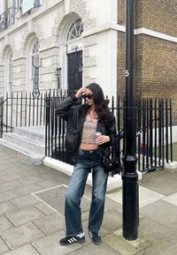 Μαύρο bomber jacket, τοπ σε λεοπάρ σχέδιο, φαρδιά μπλε τζιν, μαύρα adidas sneakers. Κρατάει ένα αναψυκτικό κοντά σε ένα τούβλινο κτίριο και σιδερένια κιγκλιδώματα.